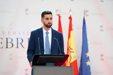 Un hombre en traje de negocios se encuentra de pie detrás de un podio con banderas al fondo.