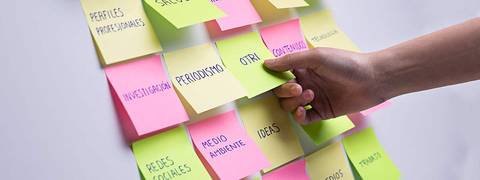 Una mano interactuando con notas adhesivas de colores que contienen palabras clave relacionadas con el periodismo y la investigación.