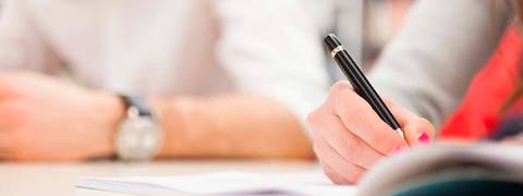 Una mano escribe con un bolígrafo sobre un cuaderno mientras se observan otras manos en la mesa.