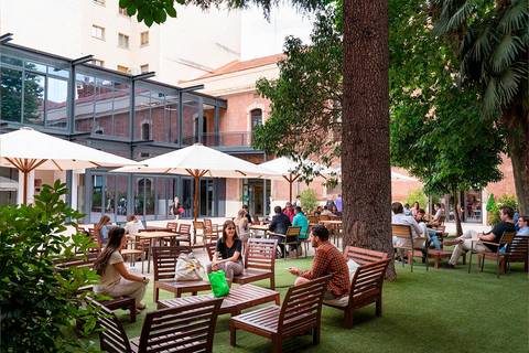 El lugar muestra un jardín con mesas y sillas, donde varias personas están conversando y disfrutando del ambiente al aire libre.