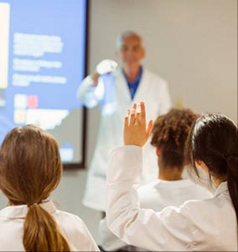 Un grupo de estudiantes en bata blanca levantan la mano durante una clase impartida por un profesor en un aula.