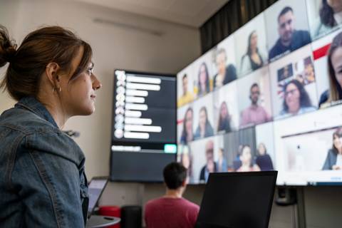 Una mujer está mirando una pantalla grande con muchas personas en videoconferencia mientras trabaja en su ordenador portátil.