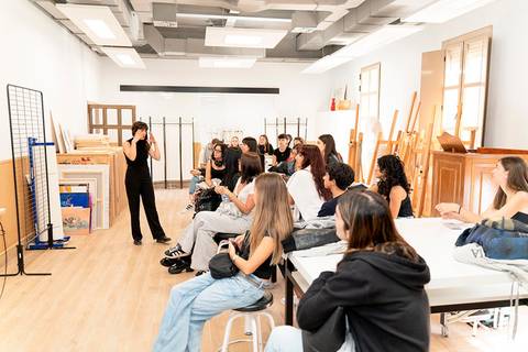 Una profesora está dando una clase a un grupo de estudiantes en un aula con mesas y sillas.