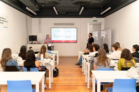 Una mujer presenta información frente a un grupo de estudiantes en un aula moderna.