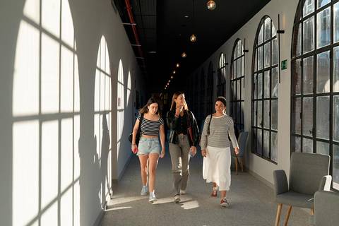 Tres mujeres caminan por un pasillo iluminado, acompañado de sombras en la pared.