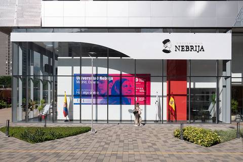La imagen muestra la entrada de la Universidad Nebrija en Madrid, con pantallas y banderas visibles.