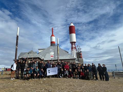 Un grupo grande de personas se encuentra posando frente a un edificio con chimeneas rojas y blancas bajo un cielo nublado.