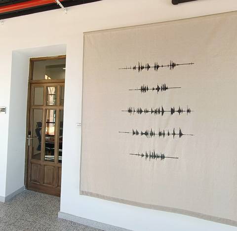 La imagen muestra una pared con una obra de arte en tela que representa ondas sonoras en color negro sobre un fondo claro.