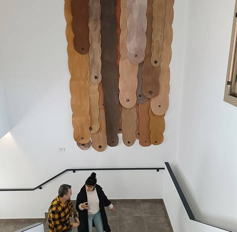 La imagen muestra una escalera con una interacción entre dos personas y una obra artística en la pared de arriba hecha de formas de colores terracota y beige.