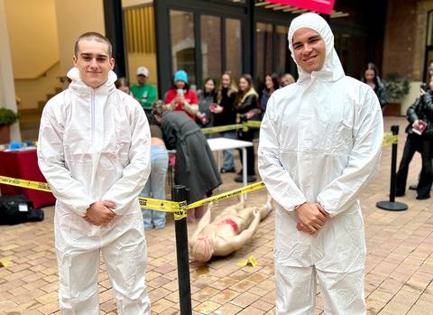 Dos personas vestidas con trajes de protección están de pie frente a un grupo de espectadores en un evento al aire libre.