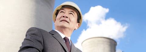 Un hombre con casco y traje de negocios observa las torres de enfriamiento de una planta industrial bajo un cielo despejado.