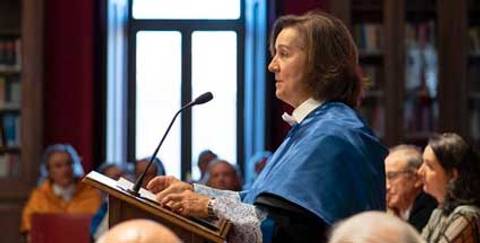 Una mujer con capa azul habla en un podio frente a una audiencia en una sala con estanterías de libros.