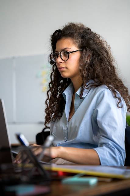 Una joven con cabello rizado y gafas trabaja concentrada frente a una computadora portátil.