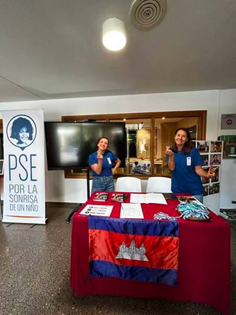 Dos mujeres sonrientes posan junto a una mesa decorada con una bandera y folletos en un evento benéfico.