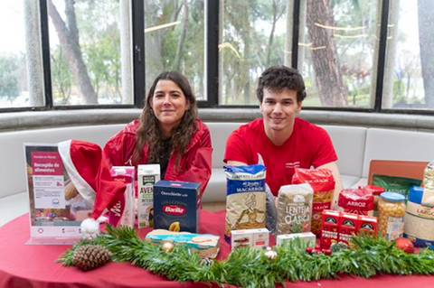 Un par de personas, una mujer y un hombre, posan junto a una mesa decorada con alimentos y carteles para una colecta de alimentos.