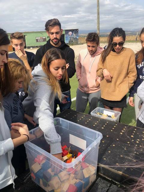 Un grupo de jóvenes observa atentamente mientras una chica explora una caja llena de bloques de colores en una actividad al aire libre.