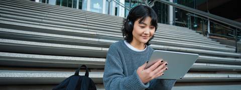Una joven con auriculares sonríe mientras utiliza una tablet sentada en unas escaleras.
