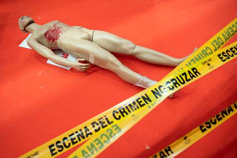 Un maniquí yace en el suelo con una marca de sangre y rodeado de cinta de emergencia que indica una escena del crimen.