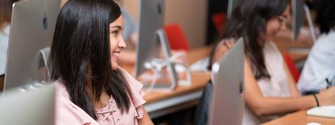 Una estudiante sonríe mientras trabaja en una computadora en un aula.