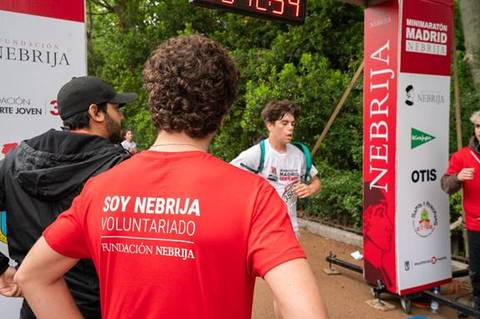 La imagen muestra a un corredor llegando a la meta en un evento deportivo con varios voluntarios de la Fundación Nebrija.