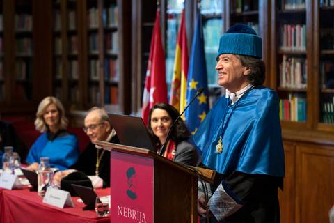 Un hombre en toga y birrete se encuentra en una ceremonia académica en una sala con estanterías de libros y banderas.