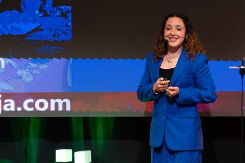 Una mujer sonriente vestida de traje azul se presenta en un evento con un fondo colorido.