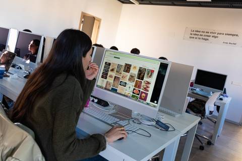 Una mujer joven está buscando imágenes en un ordenador en un aula llena de computadoras.