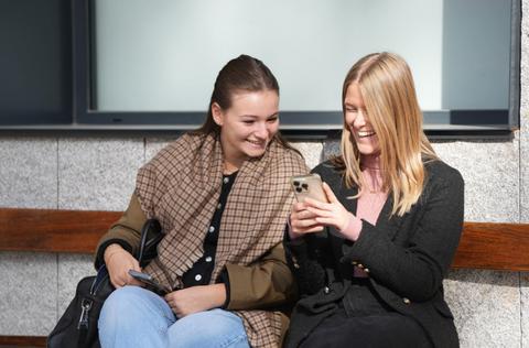 Dos jóvenes se ríen mientras miran un teléfono móvil sentadas en un banco.