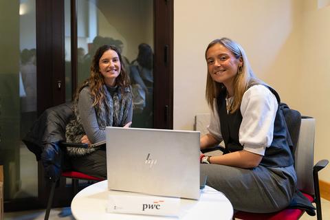 Dos mujeres sonrientes se encuentran sentadas junto a una mesa con un ordenador portátil en un ambiente de trabajo.