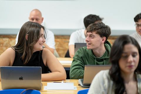 Dos estudiantes conversan en un aula con computadoras portátiles en la mesa.