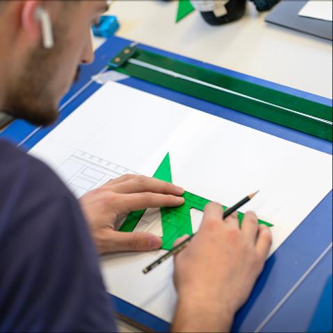 Una persona dibujando con un lápiz sobre una hoja de papel utilizando una regla verde.