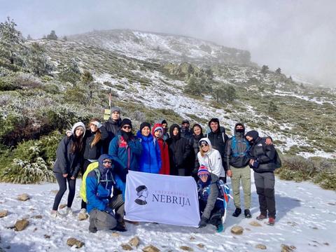 Un grupo de estudiantes de Nebrija posan en la nieve con una pancarta de su universidad en un paisaje montañoso.