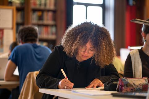 Una mujer con cabello rizado está escribiendo en una mesa dentro de una biblioteca.