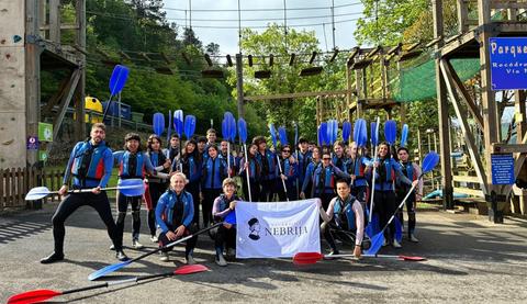 Un grupo de jóvenes uniformados con chalecos y remos posan juntos frente a un parque de aventuras al aire libre.
