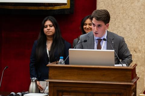 Un joven expone en un evento formal mientras dos mujeres lo observan desde el fondo.