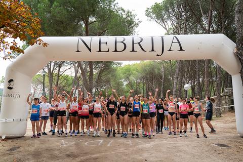 Un grupo de corredoras sonrientes se prepara para una carrera bajo un arco inflable que dice 'NEBRIJA'.