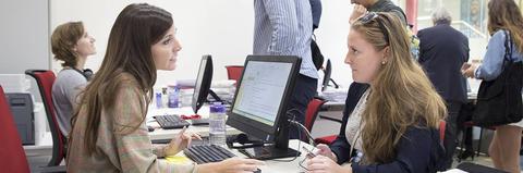 Dos mujeres se están comunicando en un entorno de oficina frente a computadoras.