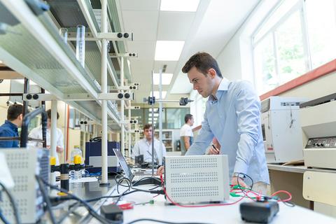 Un estudiante trabaja en un laboratorio con equipos electrónicos y otros compañeros al fondo.