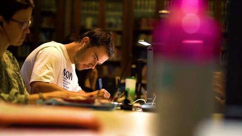 Un joven estudia concentrado en una biblioteca mientras una mujer se encuentra al fondo trabajando en su computadora.