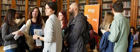 Un grupo de personas conversando en una biblioteca con un fondo de estanterías llenas de libros y un cartel de RTVE.