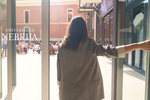 Una persona entra en un edificio universitario acompañado de un ambiente soleado y un grupo de gente en el exterior.