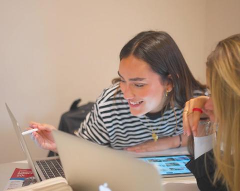 Dos mujeres se ríen y comparten ideas mientras miran una computadora portátil en una mesa.